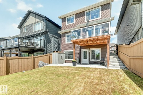 Multi-level backyard featuring a brown-toned siding exterior, a ground-level patio with a white door, and an upper-level deck with glass railings and a wood-slat underside - 2534 Wonnacott Loop, Edmonton, AB 