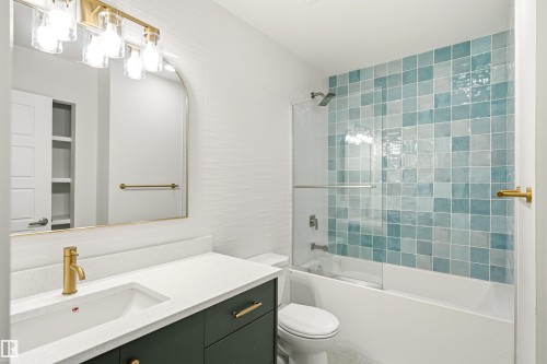 Bathroom featuring a tiled shower/tub combination with clear glass enclosure, a vanity with a white countertop and dark green cabinetry, a gold-framed arched mirror, and brass-finish fixtures - 18217 84 Avenue Nw, Edmonton, AB 