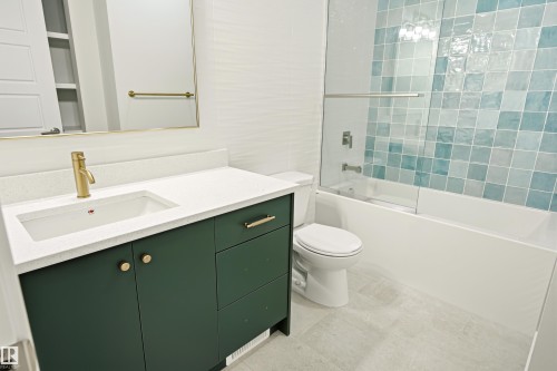 Bathroom featuring a dark green vanity with gold-tone hardware, white countertop with an integrated sink, gold-tone faucet, and a shower-over-tub with blue and white tiled surround behind a glass enclosure - 18217 84 Avenue Nw, Edmonton, AB 