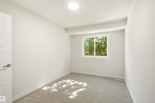 Room featuring a window with green foliage outside, light gray wall paint, gray carpeting, a paneled interior door, and a flush-mount ceiling light fixture - 18217 84 Avenue Nw, Edmonton, AB 