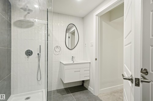 Bathroom featuring a glass-enclosed shower with a rainfall shower head and handheld sprayer, white vertical subway tile, a floating vanity with a white countertop and integrated sink, and an organic-shaped black-framed mirror - 18217 84 Avenue Nw, Edmonton, AB 