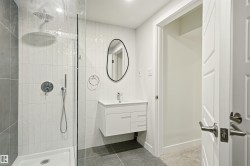 Bathroom featuring a glass-enclosed shower with a rainfall shower head and handheld sprayer, white vertical subway tile, a floating vanity with a white countertop and integrated sink, and an organic-shaped black-framed mirror - 