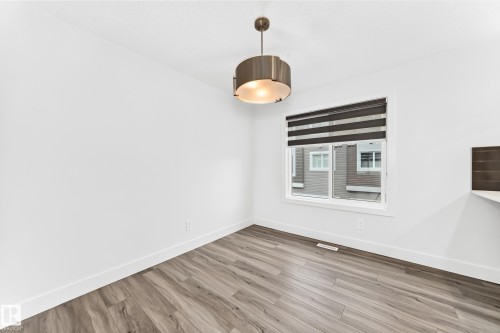 Spacious room featuring wood-finish flooring, a contemporary drum pendant light fixture, and a large window with dual-layer blinds - 17 4137 76 Street, Edmonton, AB 