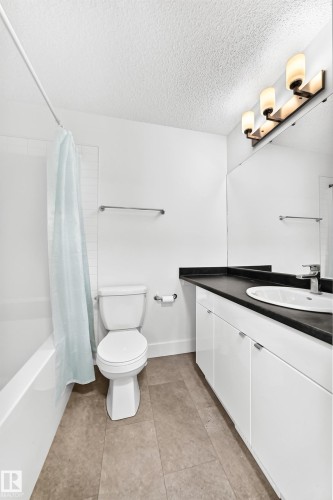 Bathroom featuring a white bathtub with tile surround, a white toilet, a vanity with white cabinetry, dark countertop, and an integrated sink, and square tile flooring - 17 4137 76 Street, Edmonton, AB 