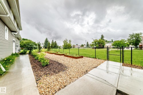 Exterior siding in a neutral tone with white trim, concrete walkway, gravel landscaping beds, multiple raised garden beds, and a black chain-link fence bordering a grassy area - 17 4137 76 Street, Edmonton, AB 