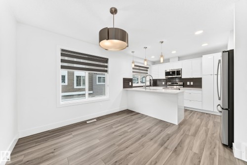 Open-concept kitchen and dining area featuring wood-finish flooring, white cabinetry, stainless steel appliances, a center island with sink, and modern pendant lighting - 17 4137 76 Street, Edmonton, AB 