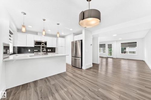 Modern kitchen featuring white cabinetry, stainless steel appliances, a large island with white countertops, a dark subway tile backsplash, and wood-finish flooring - 17 4137 76 Street, Edmonton, AB 