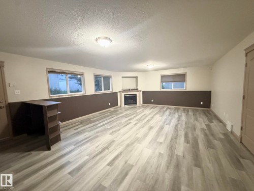 Spacious room featuring light wood-finish flooring, a built-in electric fireplace with a white mantel, and multiple windows with roller blinds - 1978 Tomlinson Green Green, Edmonton, AB 
