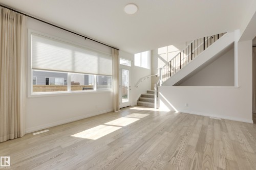 Open concept living space featuring light wood-finish flooring, a contemporary staircase with metal spindles, and a multi-pane glass door - 3249 Dixon Way, Edmonton, AB 
