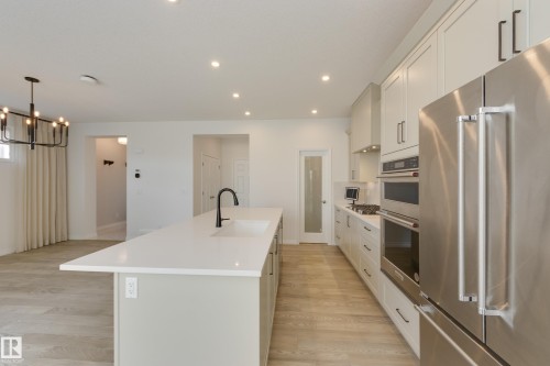 Modern kitchen featuring a large island with an undermount sink and black faucet, light wood-finish flooring, recessed lighting, white shaker cabinetry with dark hardware, and stainless steel appliances including a built-in oven and refrigerator - 3249 Dixon Way, Edmonton, AB 