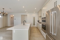 Modern kitchen featuring a large island with an undermount sink and black faucet, light wood-finish flooring, recessed lighting, white shaker cabinetry with dark hardware, and stainless steel appliances including a built-in oven and refrigerator - 