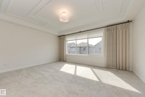 Spacious room featuring a coffered ceiling, a large window with roller shades and drapery, neutral carpeting, and a semi-flush mount ceiling fixture - 3249 Dixon Way, Edmonton, AB 