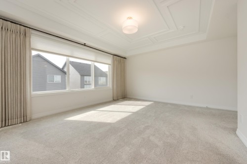 Spacious room featuring light-colored carpeting, a large window with roller shades and drapery, a decorative coffered ceiling, and a flush-mount light fixture - 3249 Dixon Way, Edmonton, AB 