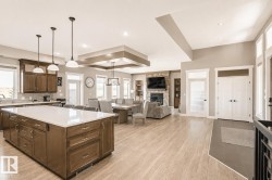 Open-concept living area with wood-finish flooring, a stone-faced fireplace, a kitchen island with a light-toned countertop, recessed ceiling lights, and multiple windows providing ambient light - 