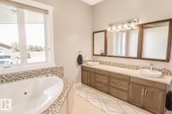 Bathroom featuring a large soaking tub with mosaic tile surround, dual vessel sinks, a wide vanity with wood-finish cabinetry, and a multi-light fixture over three framed mirrors - 