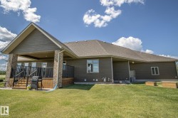 Expansive rear exterior featuring a covered deck, stone-accented pillars, dark horizontal siding, a pitched roofline, and a manicured lawn - 