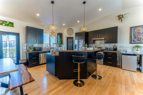 Open-concept kitchen featuring dark cabinetry, stainless steel appliances, a central island with bar seating, wood-finish flooring, and a subway tile backsplash - 4804 64 Avenue, Cold Lake, AB 