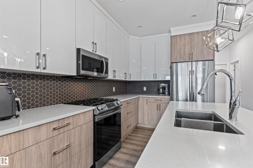 Contemporary kitchen featuring two-tone cabinetry, a dark hexagon tile backsplash, light stone countertops, stainless steel appliances, and a geometric pendant light fixture - 8046 Kiriak Link, Edmonton, AB - Indoor Photo Showing Kitchen With Double Sink With Upgraded Kitchen
