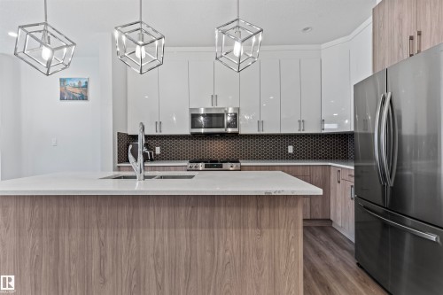 Gourmet kitchen featuring a wood-finish island with integrated sink, white countertops, stainless steel appliances, white upper cabinetry, and dark honeycomb tile backsplash - 8046 Kiriak Link, Edmonton, AB - Indoor Photo Showing Kitchen With Upgraded Kitchen