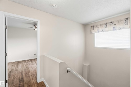 Interior hallway featuring a staircase with a white handrail, a window with horizontal blinds and a decorative valance, and a doorway opening to a room with wood-finish flooring and a ceiling fan - 3312 106 Avenue, Edmonton, AB 