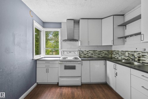 Kitchen featuring white cabinetry, dark countertops, a mosaic tile backsplash, a stainless steel range hood, and wood-finish flooring - 7520 106 Street, Edmonton, AB 