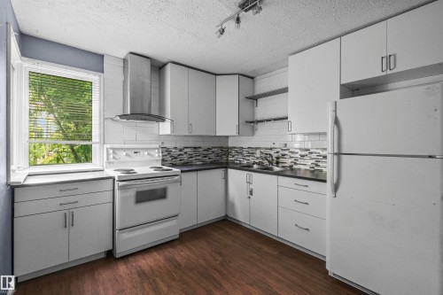 Kitchen featuring white cabinetry, dark countertops, a mosaic tile backsplash, a stainless steel range hood, and wood-finish flooring - 7520 106 Street, Edmonton, AB 