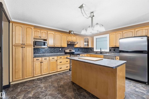 Kitchen featuring light wood cabinetry, dark mosaic tile backsplash, stainless steel appliances, and a central island with a grey countertop - 17303 107 Street, Edmonton, AB 
