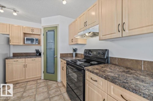 Kitchen featuring light wood-finish cabinetry, dark speckled countertops, a black electric range, a microwave, and a frosted glass pantry door - 8509 7 Avenue, Edmonton, AB 