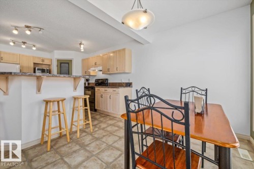 Open-concept kitchen and dining area featuring light wood cabinetry, stone-finish countertops, tiled flooring, track lighting, and a breakfast bar - 8509 7 Avenue, Edmonton, AB 
