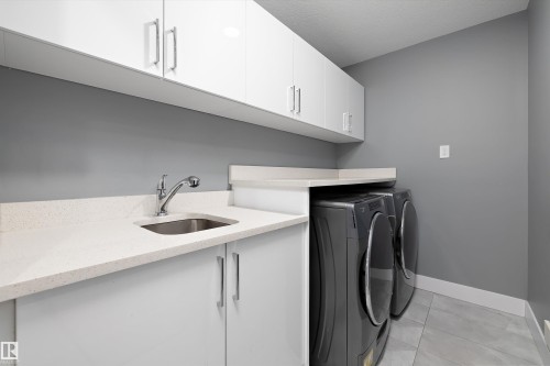 Dedicated laundry area featuring white upper and lower cabinetry, a stainless steel utility sink, and light-toned countertops - 6084 179 Avenue, Edmonton, AB 
