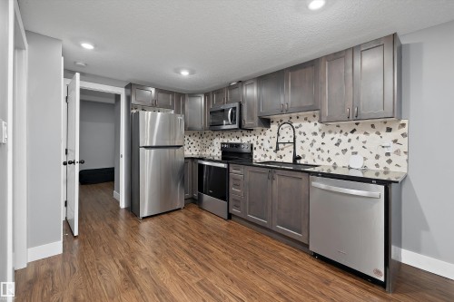 Kitchen featuring wood-finish flooring, gray cabinetry, stainless steel appliances, a black countertop, and a decorative tile backsplash - 6084 179 Avenue, Edmonton, AB 