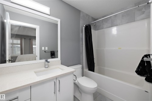 Ensuite bathroom featuring a rectangular undermount sink, a light-toned countertop, white cabinetry with silver-toned hardware, a framed mirror, and a shower-tub combination with light gray wall tiling - 6084 179 Avenue, Edmonton, AB 