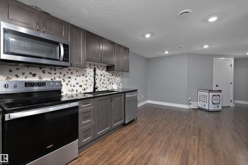 Modern kitchen featuring stainless steel appliances, dark wood-finish cabinetry, black countertops, and a mosaic tile backsplash - 6084 179 Avenue, Edmonton, AB 