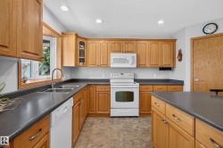 Bright kitchen featuring light wood cabinetry, dark countertops, a double basin stainless steel sink, and white appliances - 