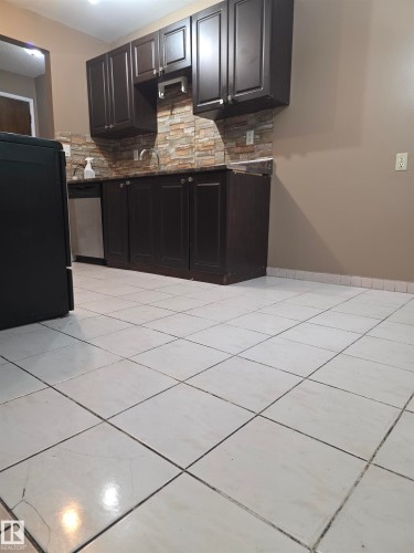 Kitchen featuring dark wood-finish cabinetry, stainless steel dishwasher, stone tile backsplash, and light-colored tile flooring - 307 11907 81 Street Nw, Edmonton, AB 