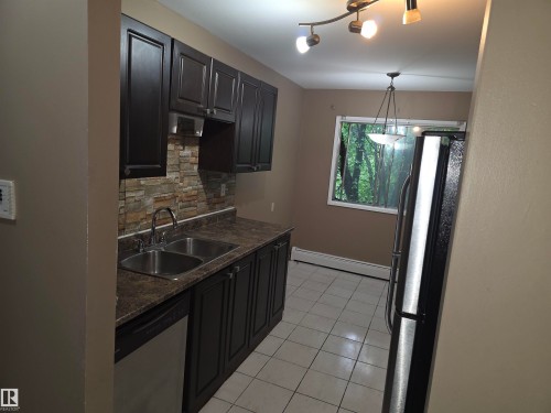 Kitchen featuring dark cabinetry, stone-look backsplash, dual basin stainless steel sink, and light tile flooring - 307 11907 81 Street Nw, Edmonton, AB 