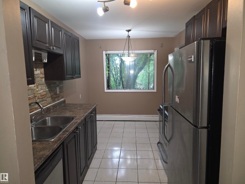 Kitchen featuring dark wood-finish cabinetry, a double basin stainless steel sink, and a stacked stone backsplash - 307 11907 81 Street Nw, Edmonton, AB 