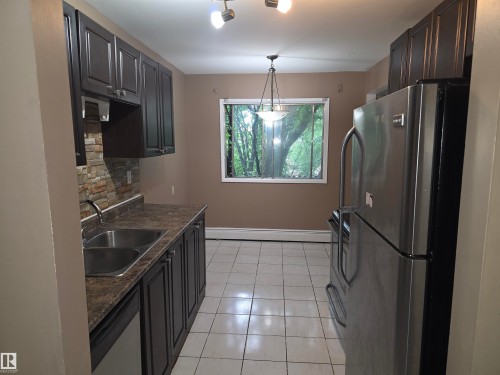 Kitchen featuring dark wood-finish cabinetry, a stone-look backsplash, a dual basin stainless steel sink, and a stainless steel refrigerator - 307 11907 81 Street Nw, Edmonton, AB 