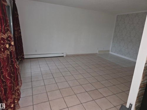 Spacious room featuring light-toned square tile flooring, white baseboards, and a feature wall with patterned wallpaper - 307 11907 81 Street Nw, Edmonton, AB 