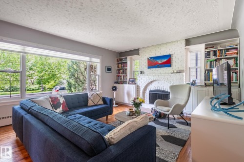 Living area featuring hardwood flooring, a large picture window, a white brick fireplace with an arched opening, and built-in shelving with cabinetry - 11634 74 Avenue Nw, Edmonton, AB 