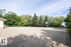 Expansive paved surface featuring a basketball hoop, bordered by mature trees and a section of a light-colored building - 