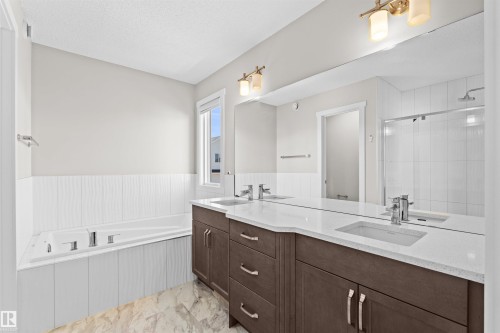 Contemporary bathroom featuring a spacious built-in tub, dual vanities with integrated sinks, a large wall-mounted mirror, a glass-enclosed shower, and wood-finish cabinetry - 4673 Alwood Way, Edmonton, AB 