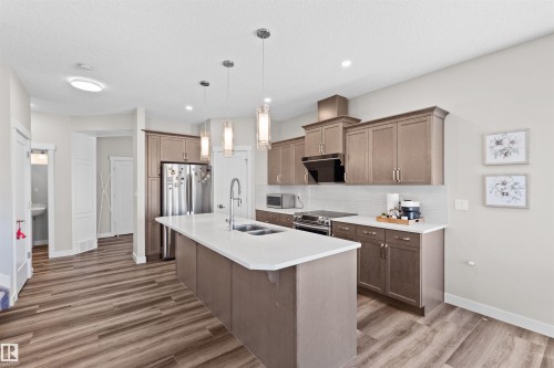 Modern kitchen featuring an expansive center island with an undermount sink, pendant lighting, wood-finish flooring, and shaker-style cabinetry with stainless steel appliances - 4673 Alwood Way, Edmonton, AB 