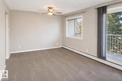 Neutral tone walls, carpeted flooring, a ceiling fan with integrated lighting, a large window, and a glass sliding door with balcony access - #223 5125 Riverbend Rd Nw, Edmonton, AB - Indoor Photo Showing Other Room