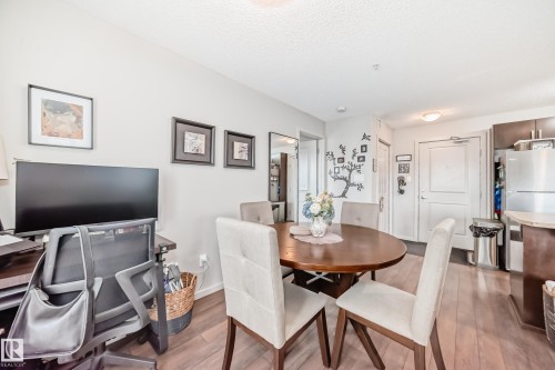 Open-concept living area with wood-finish flooring, featuring a dedicated desk area, a dining space with an oval table, and a partial view of the kitchen with dark cabinetry and stainless steel appliances - 311 142 Ebbers Boulevard, Edmonton, AB 