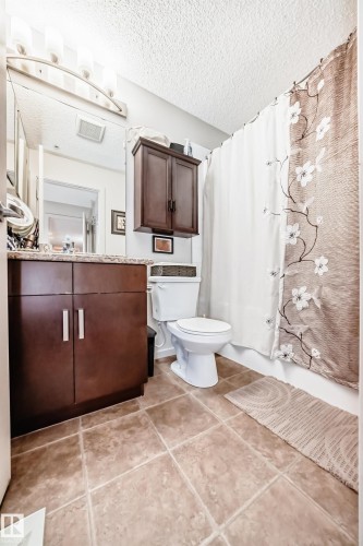Bathroom featuring a dark wood-finish vanity with a stone-finish countertop, a wall-mounted medicine cabinet, a toilet, and tiled flooring - 311 142 Ebbers Boulevard, Edmonton, AB 