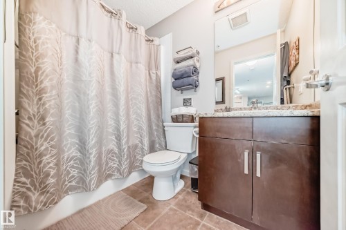 Full bathroom featuring a dark wood-finish vanity with a light-toned countertop, a rectangular mirror, a white toilet, and textured floor tiling - 311 142 Ebbers Boulevard, Edmonton, AB 