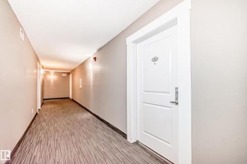 Corridor featuring light gray walls, textured patterned carpeting, and white trim, leading to a white paneled door with silver-tone hardware - 311 142 Ebbers Boulevard, Edmonton, AB 