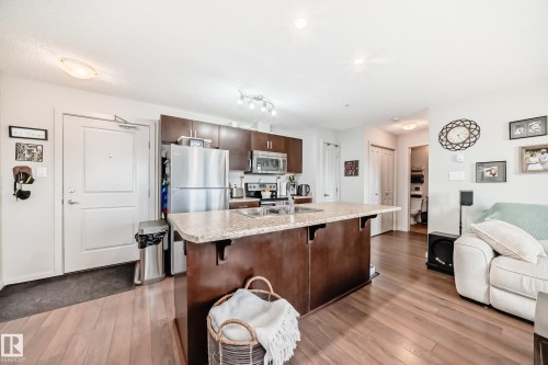 Open-concept kitchen and living area featuring wood-finish flooring, dark wood cabinetry, stainless steel appliances, and a granite-look island with an integrated sink - 311 142 Ebbers Boulevard, Edmonton, AB 