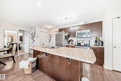 Kitchen featuring a granite-look island with an undermount sink, rich wood-finish cabinetry, and stainless steel appliances - 311 142 Ebbers Boulevard, Edmonton, AB 